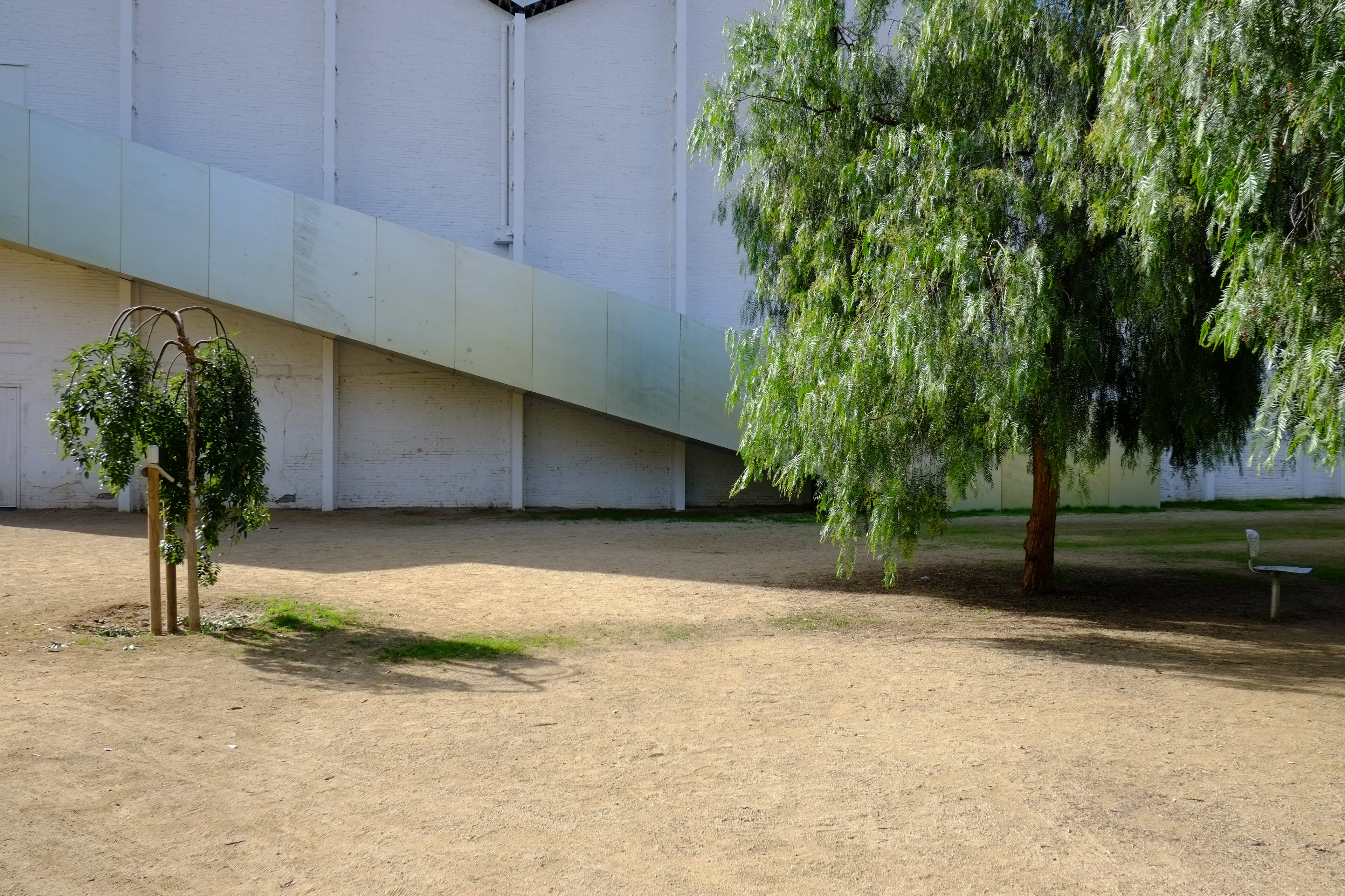 A glazed ramp cuts across a whitewashed factory wall, pepper trees in front