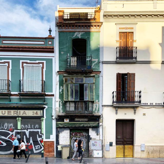 Three narrow façades on Calle Amor de Dios in Seville: a green-tiled corner shop with a graffitied shutter, a derelict green house with a glazed gallery, and a cream house with brown shutters, two women photographing the middle doorway