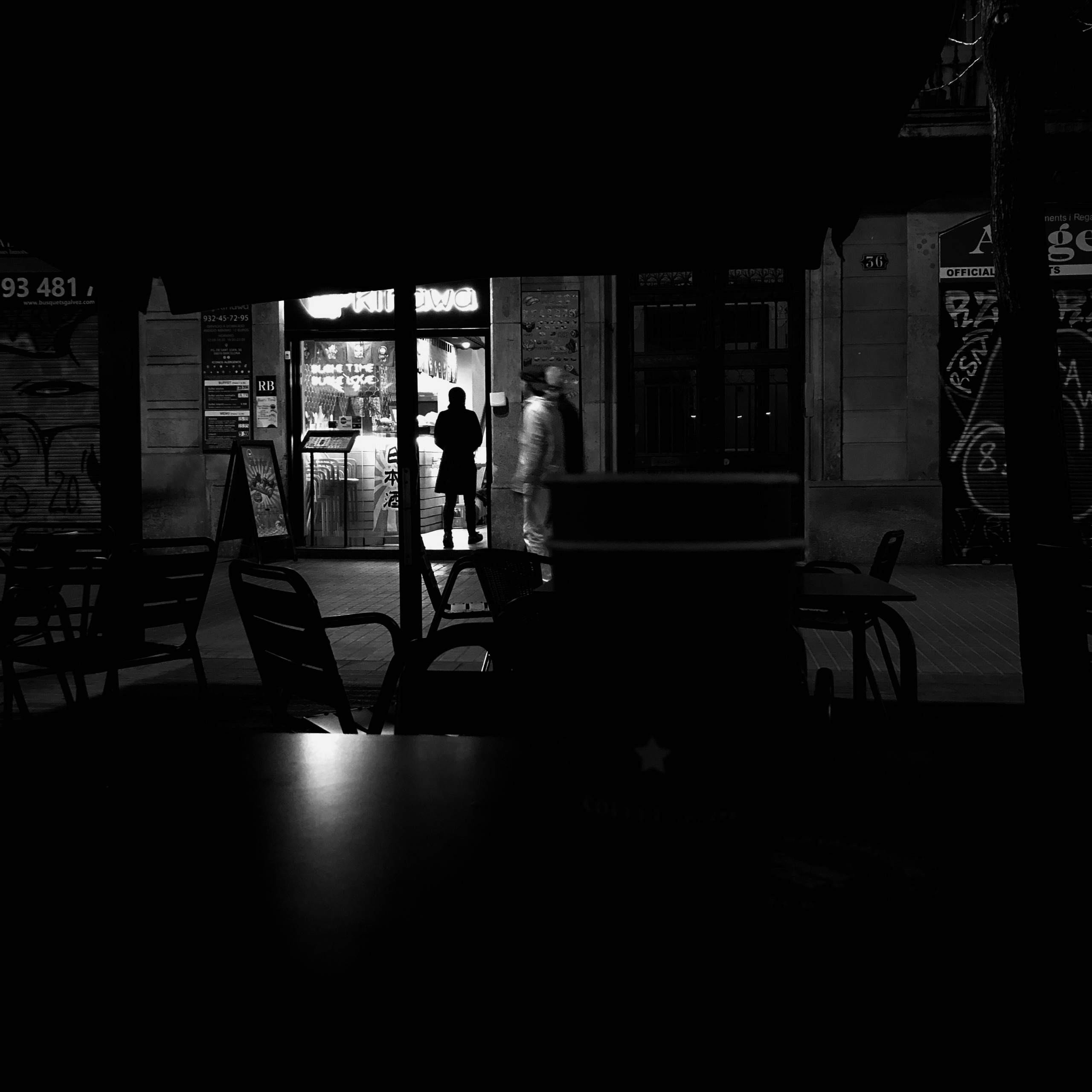From a dark cafe terrace, a lit sushi bar holds one figure at the counter while another walks on