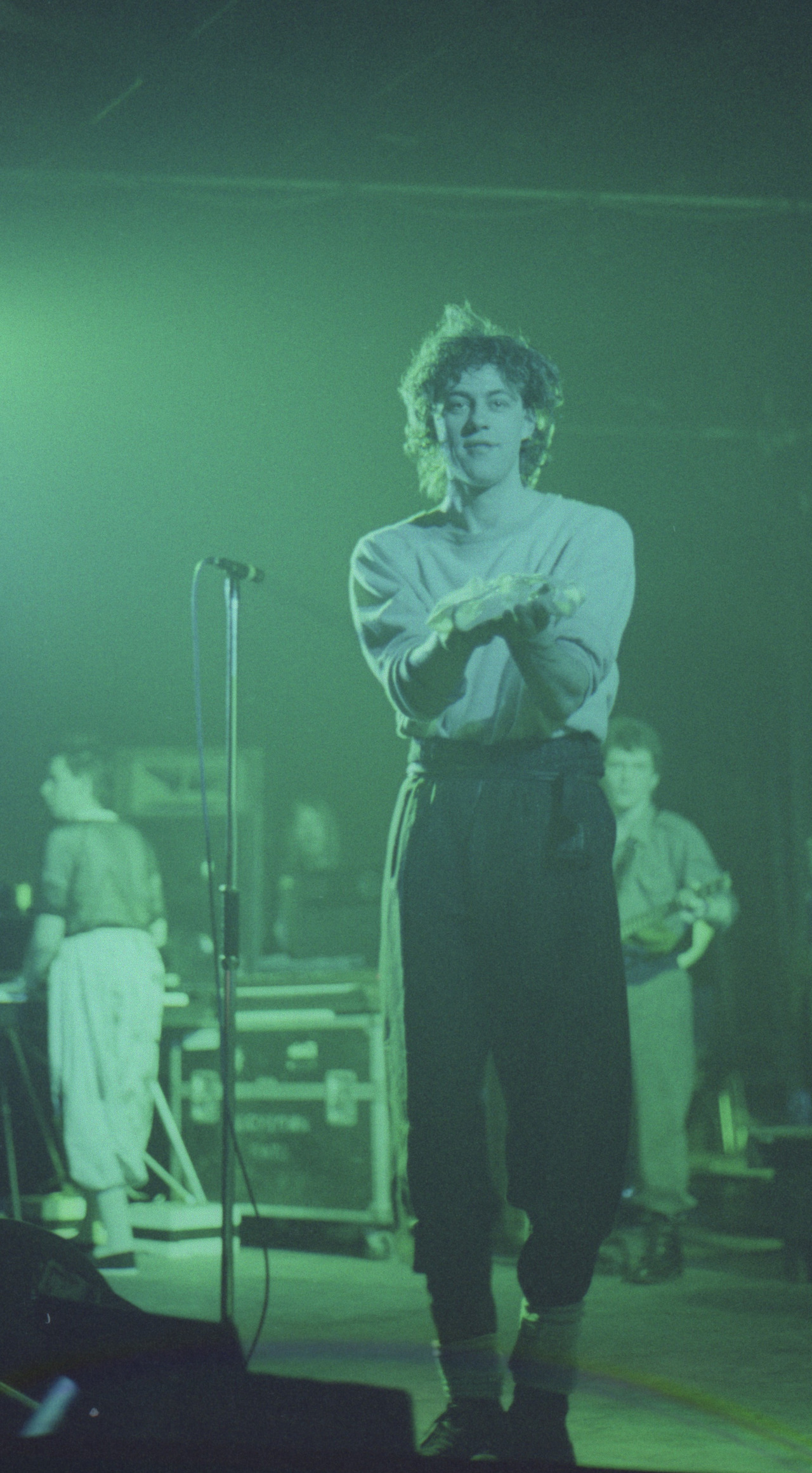 Bob Geldof of the Boomtown Rats at a mic stand in green stage light, hands clasped, keyboard player and guitarist behind him — Dundee University Student Union, 1983
