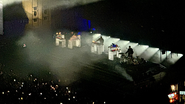 RÜFÜS DU SOL play behind caged rigs in a haze of smoke, white beams raking the stage and a crowd of phone lights below — Palau Sant Jordi, Barcelona, 2026