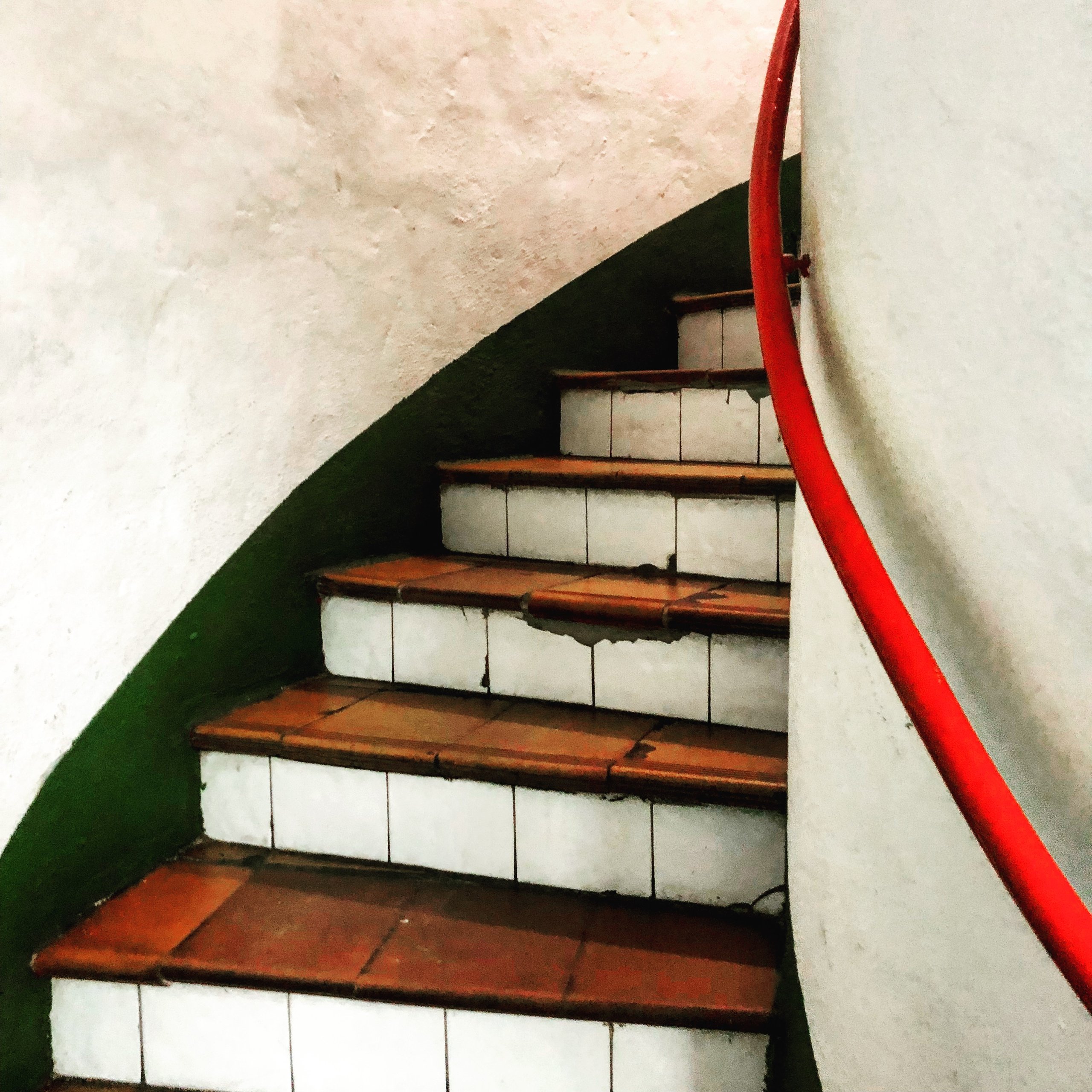 A spiral stair turns past a red handrail, terracotta treads on white tiles