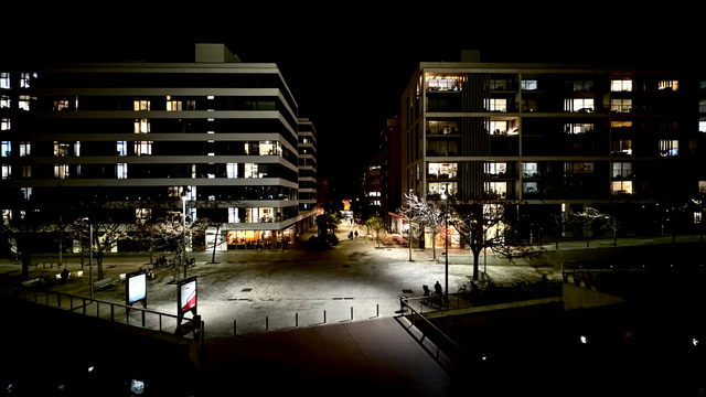 Two apartment blocks with lit windows flank a paved square at night, a café glowing at street level, bare trees and a few passers-by below