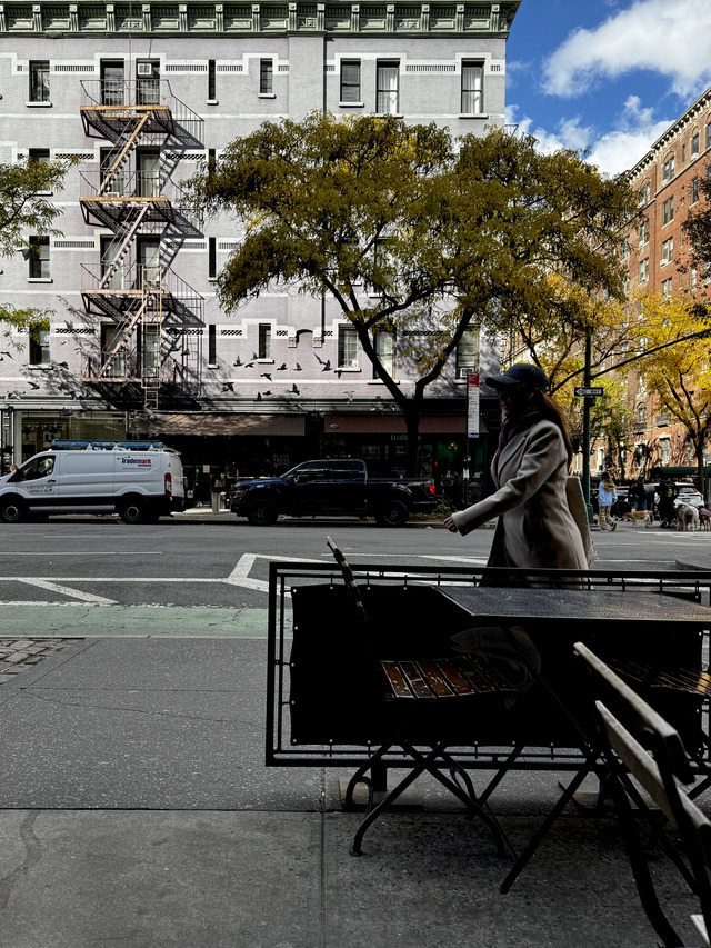 Pigeons burst past a lavender corner building as a woman crosses the street