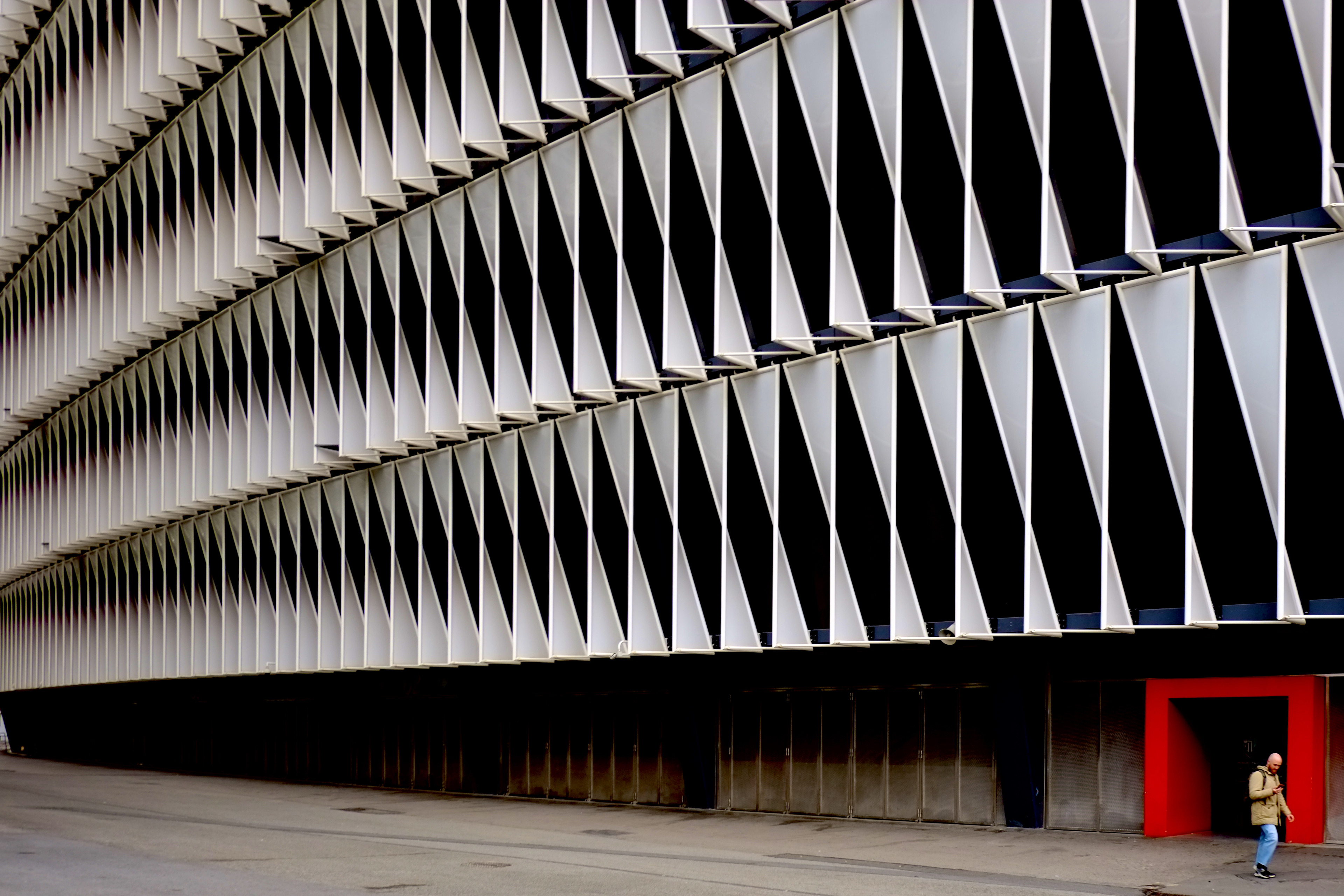 White louvres pleat a stadium facade; a passer-by leaves through a red doorway