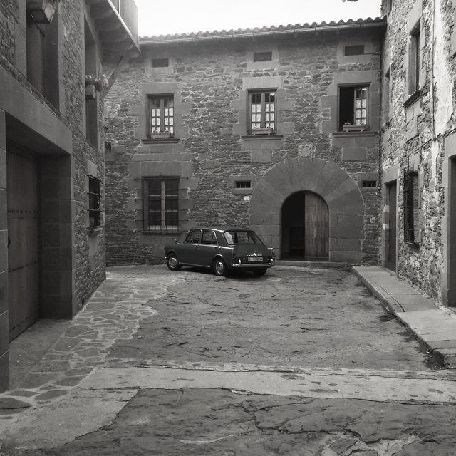 Old car parked on a worn stone square before a stone manor, monochrome