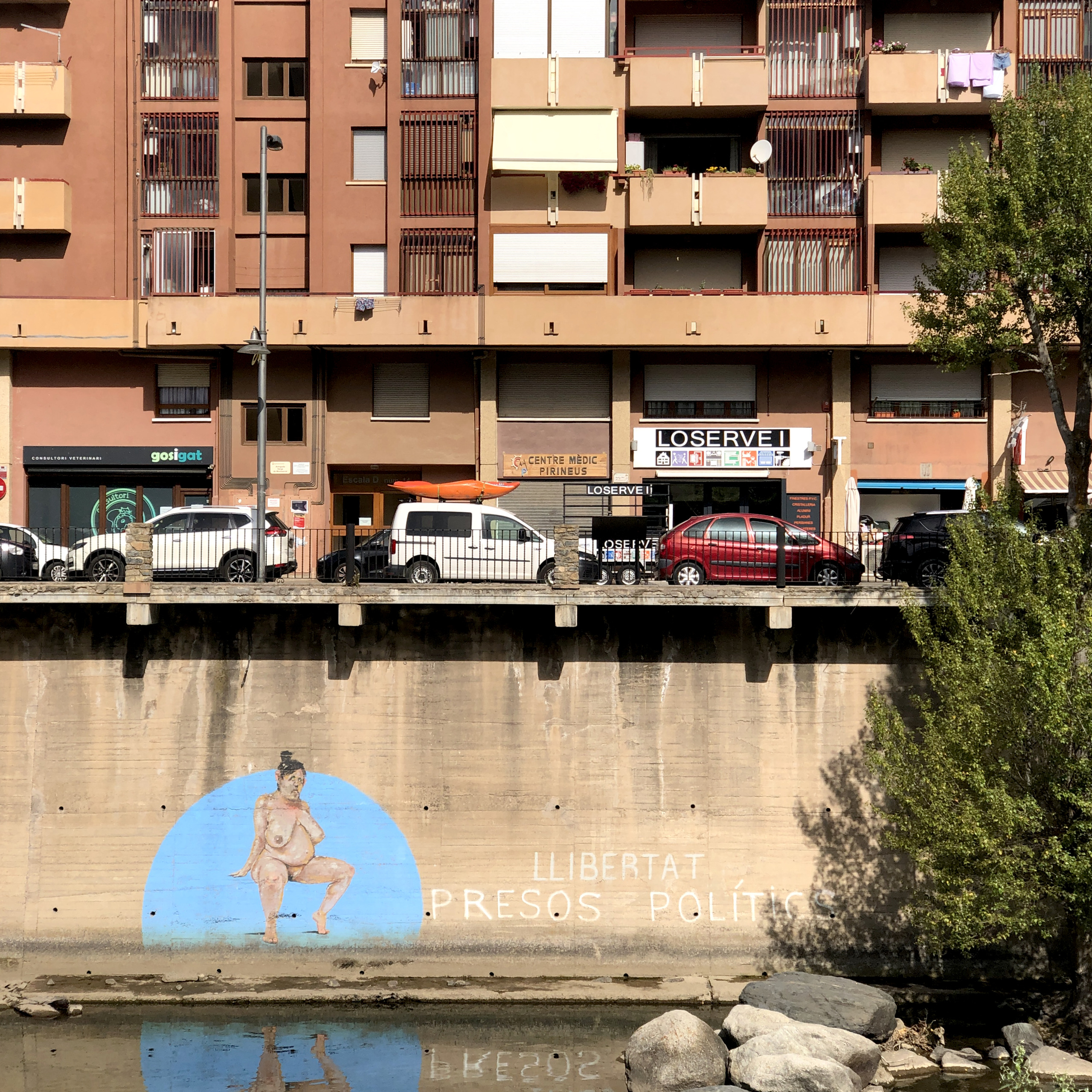 A riverside wall below apartment balconies in La Seu d'Urgell carries a mural of a seated nude figure in a blue disc beside painted words, cars parked on the embankment above