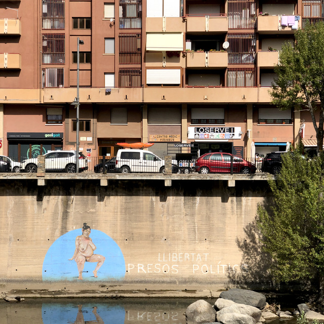 A riverside wall below apartment balconies in La Seu d'Urgell carries a mural of a seated nude figure in a blue disc beside painted words, cars parked on the embankment above