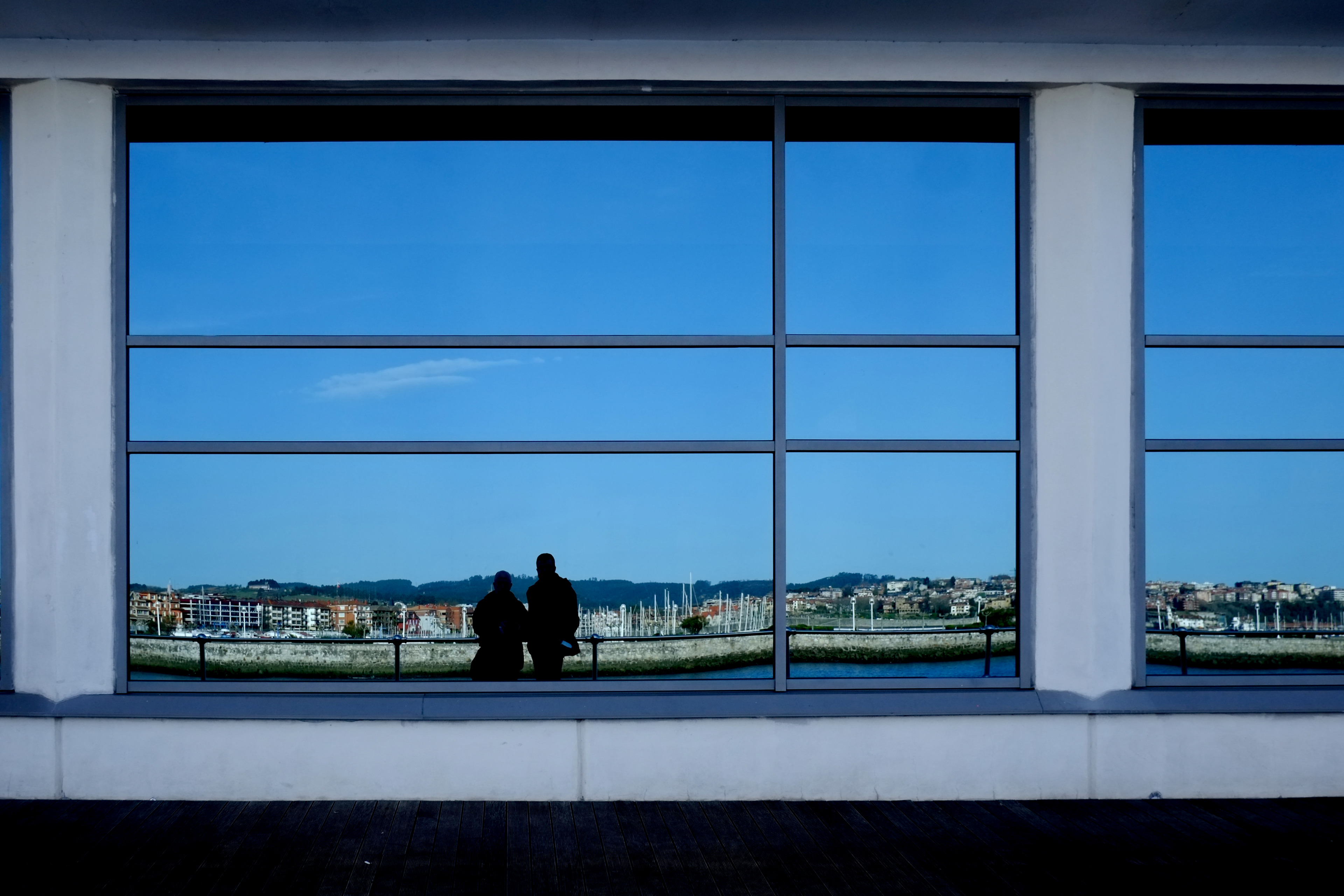 Two silhouettes at a rail, a marina town framed pane by pane in window glass