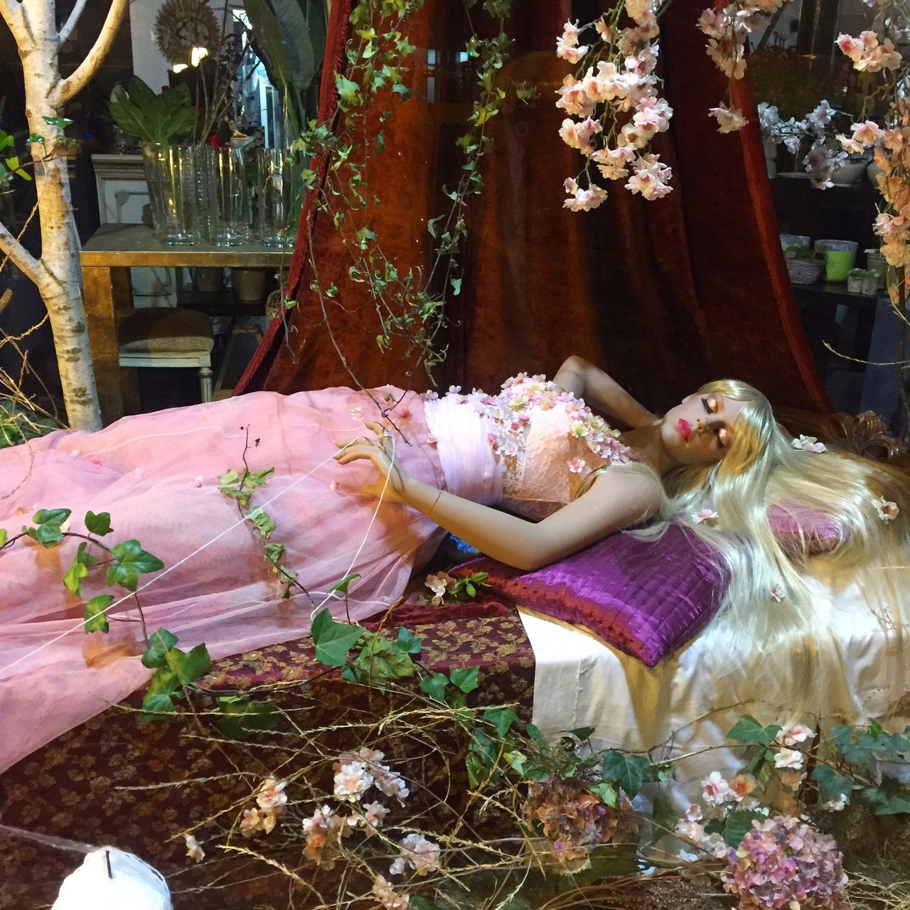 Shop-window tableau: a doll in a pink gown reclining among blossom and ivy