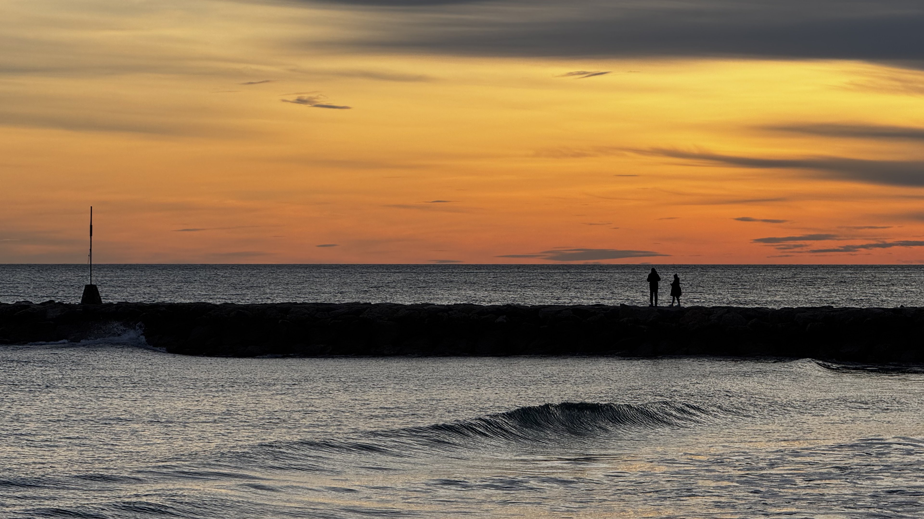 Two figures on the breakwater under an orange evening sky