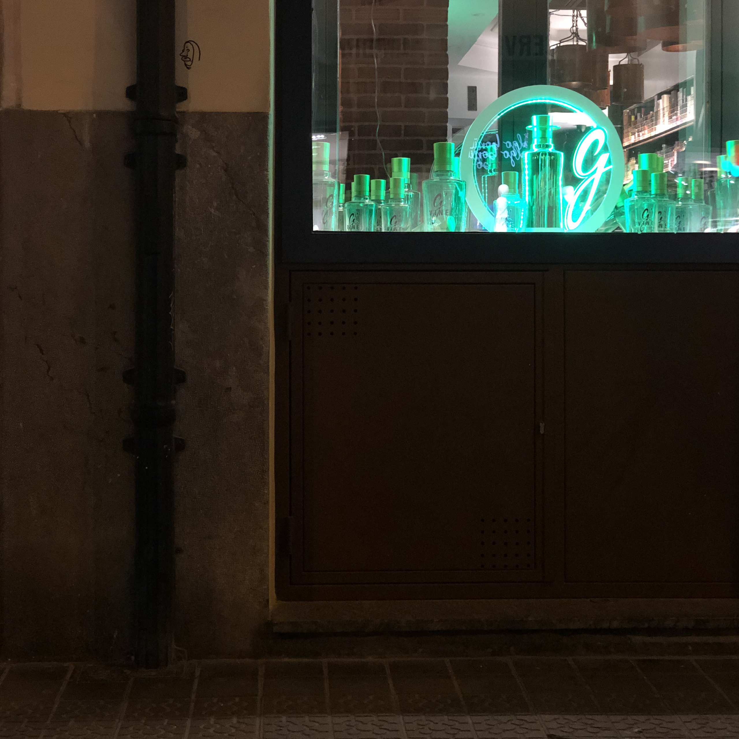 Ranks of gin bottles glow green in a dark shop window beside an old drainpipe