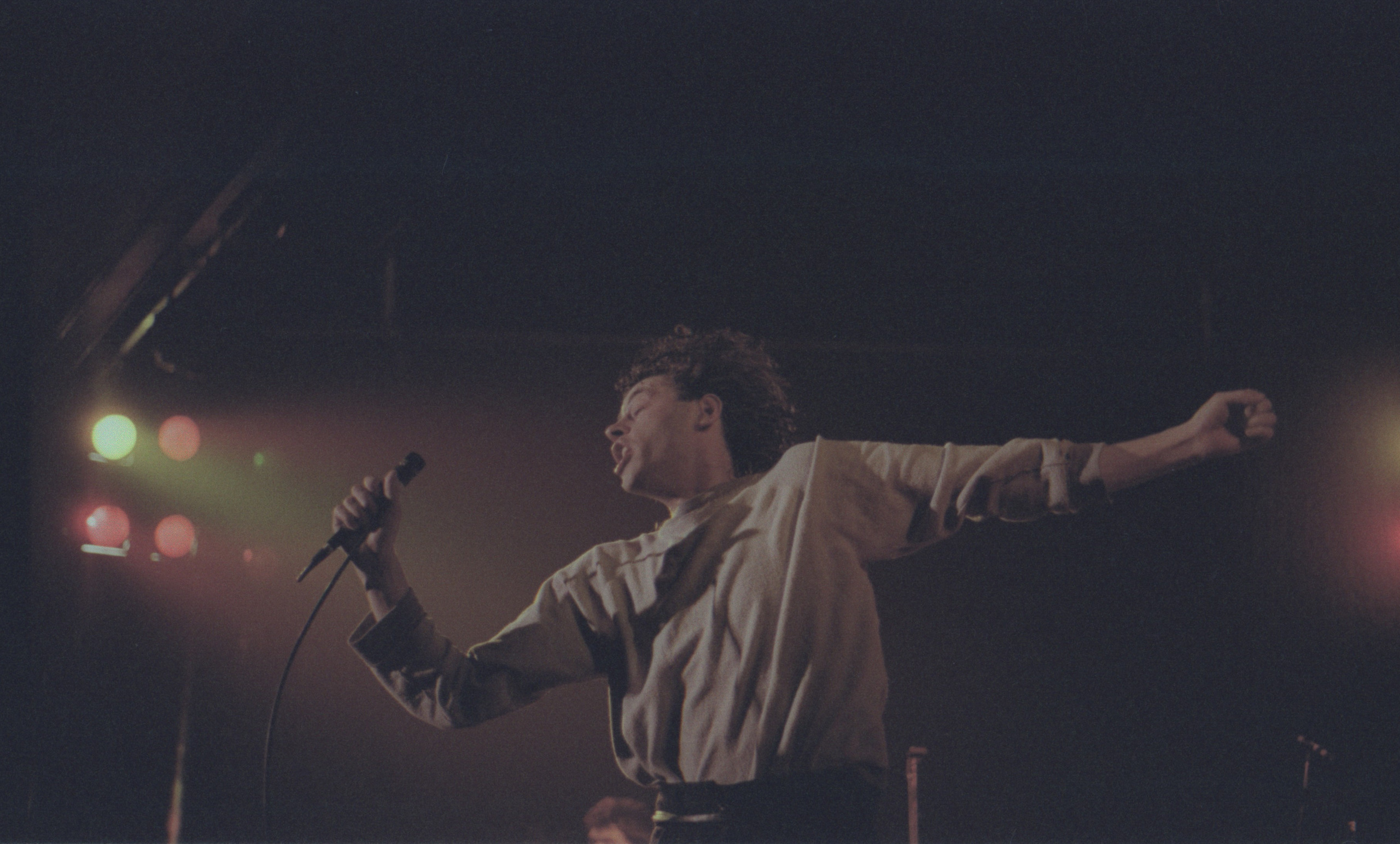 Bob Geldof of the Boomtown Rats flings both arms wide on a dark stage, microphone in one hand, coloured stage lights behind — Dundee University Student Union, 1983