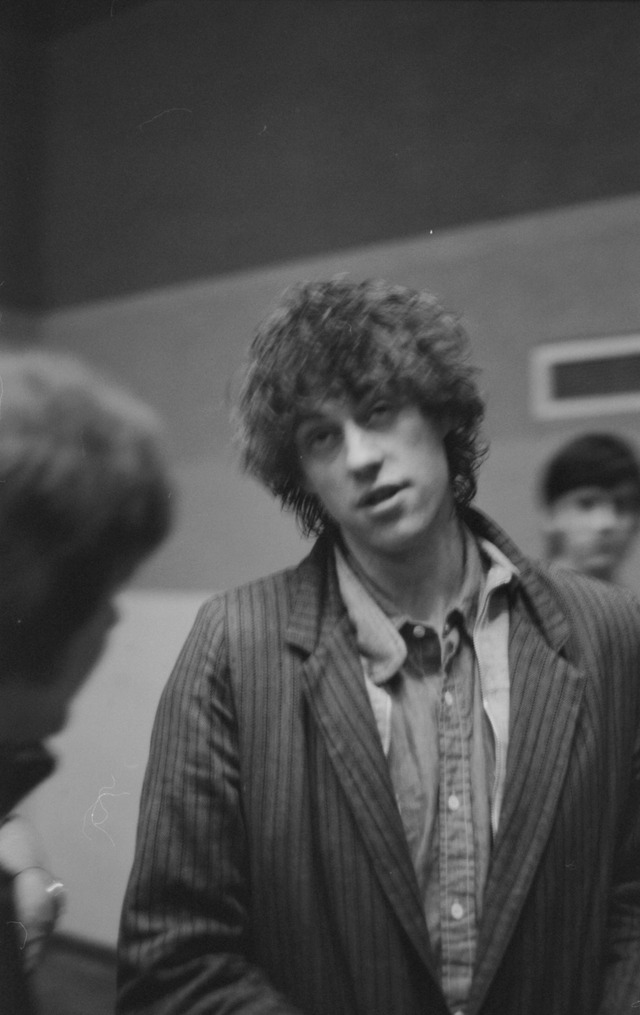 Bob Geldof of the Boomtown Rats in a pinstripe jacket, mid-sentence, over the shoulder of a listener, another face behind — Dundee University Student Union, 1983