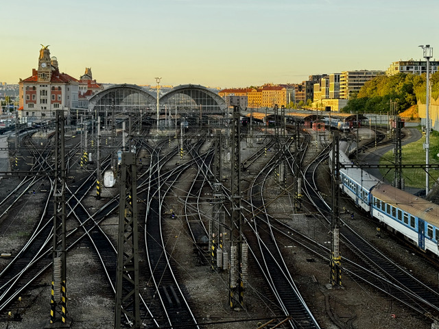 Tracks and catenary masts fan out below Prague's main station, its twin arched train sheds and clock tower lit by evening sun, a blue and white train waiting at the right