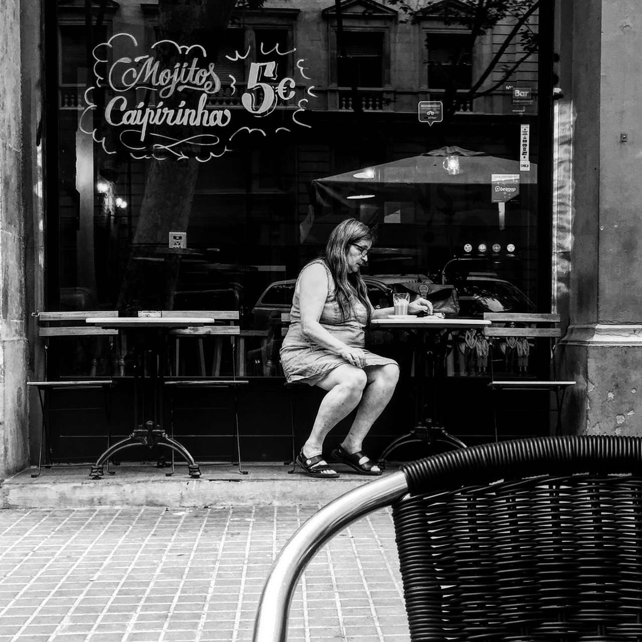 Woman seated in a café window beneath a chalked cocktail sign, monochrome