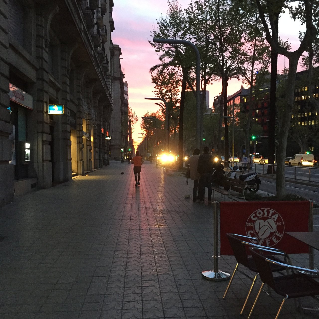 City street at first light, pink sky above lit shopfronts and bare trees